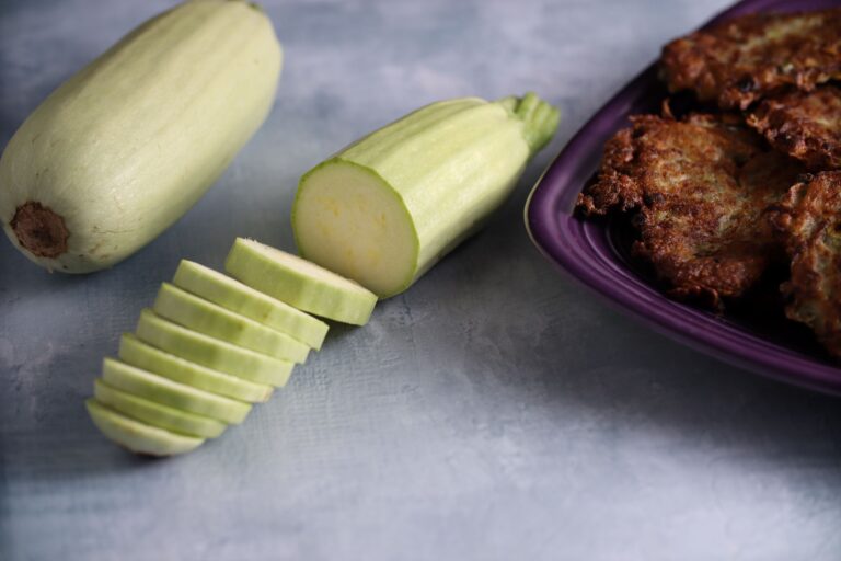 Summer Squash Seeds, Kolsuz Sakız One whole and one sliced Sakiz summer squash fruits are photographed next to a plate full of mucver (squash fritters) on photography backdrop with concrete pattern.