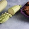 One whole and one sliced Sakiz summer squash fruits are photographed next to a plate full of mucver (squash fritters) on photography backdrop with concrete pattern.